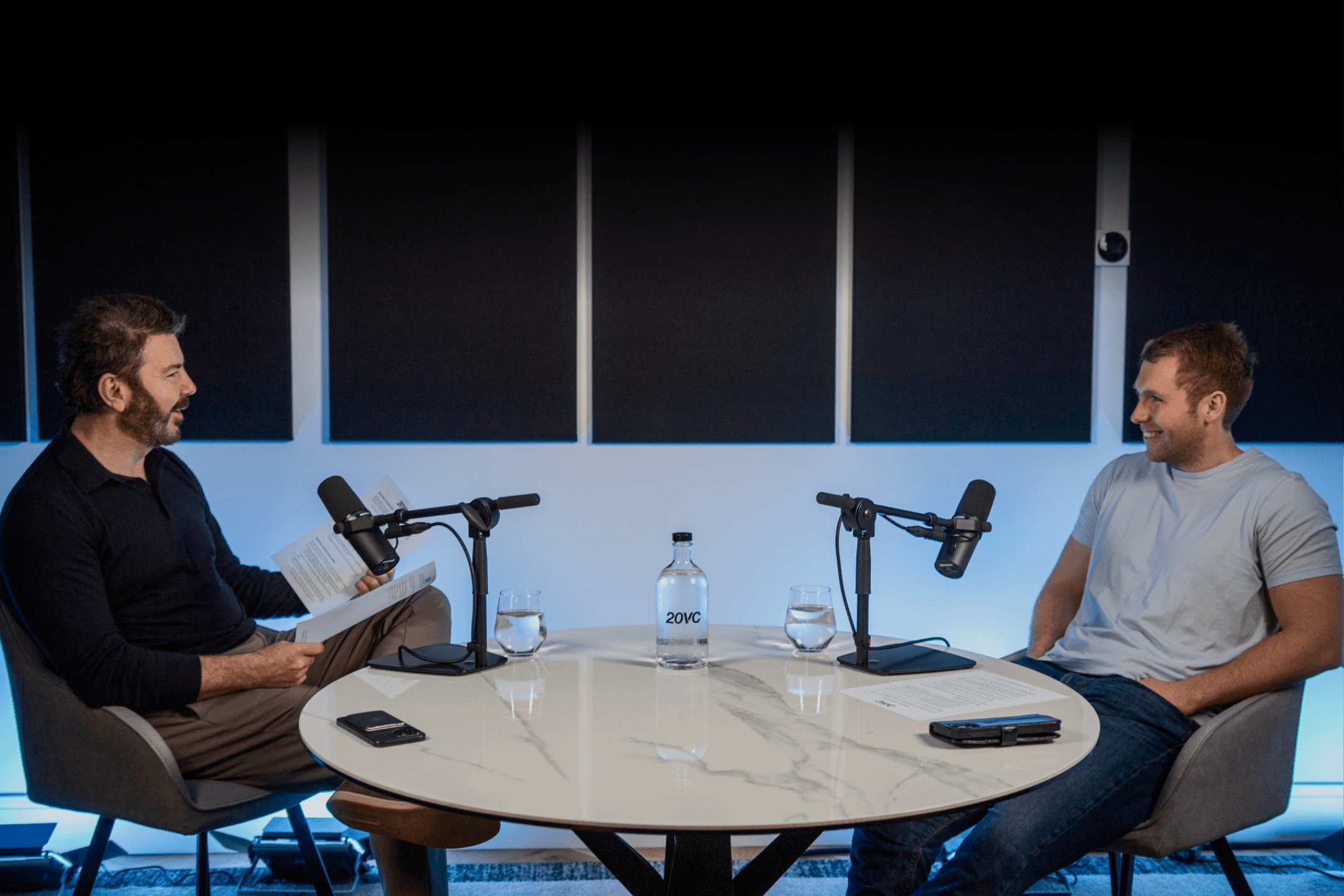 Two participants sit across microphones at a round table in the 20VC recording studio