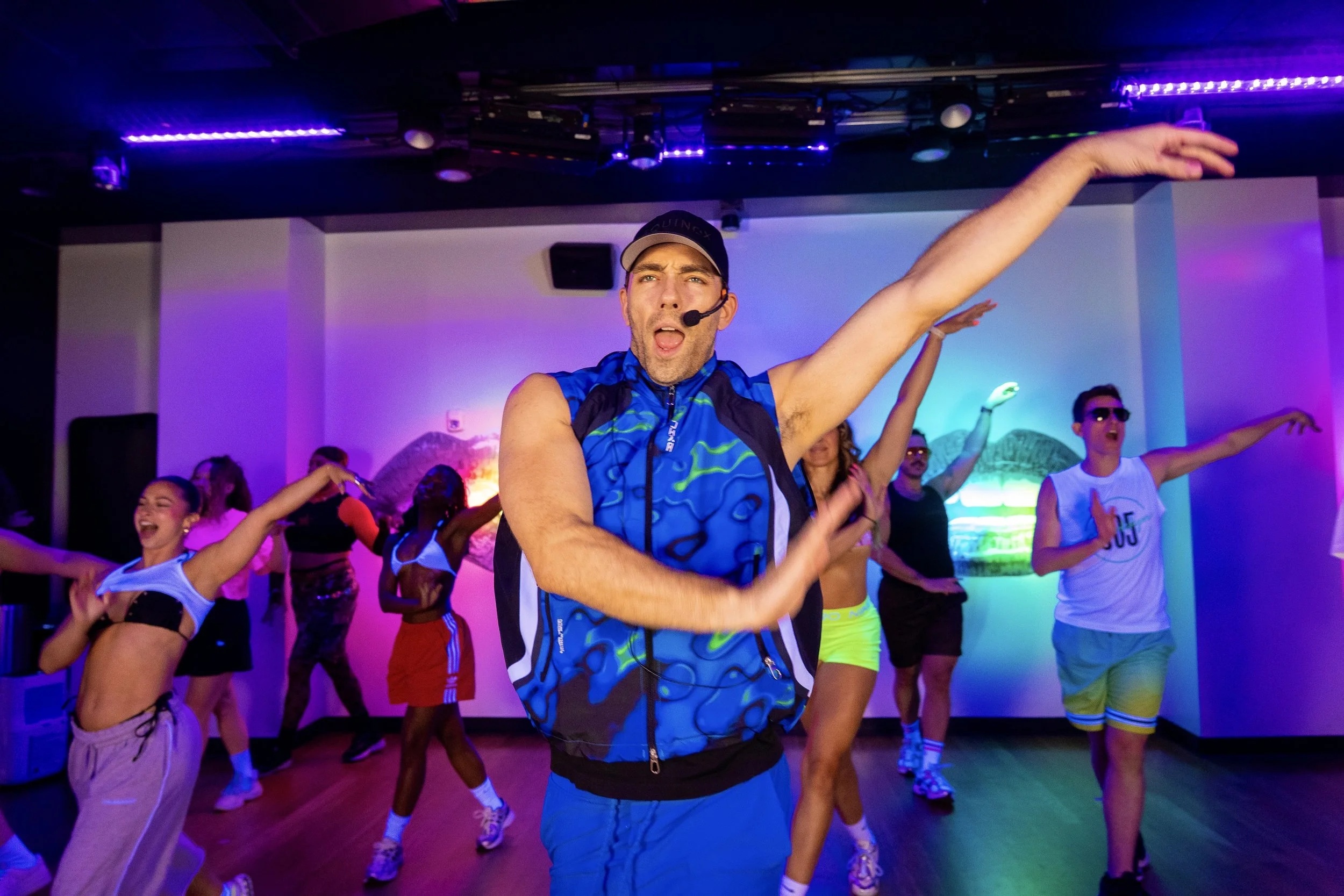 An instructor leads participants through a dance move in a purple-lit 305 Fitness studio