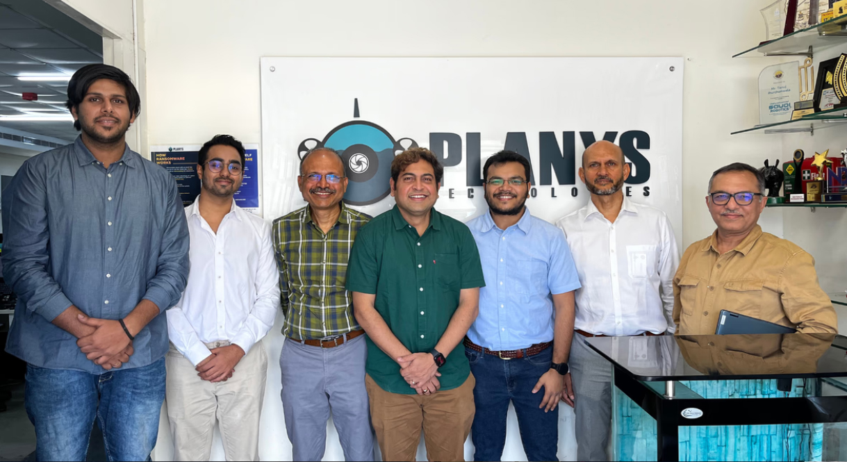 A group posing in the Planys Technologies office in front of its company sign