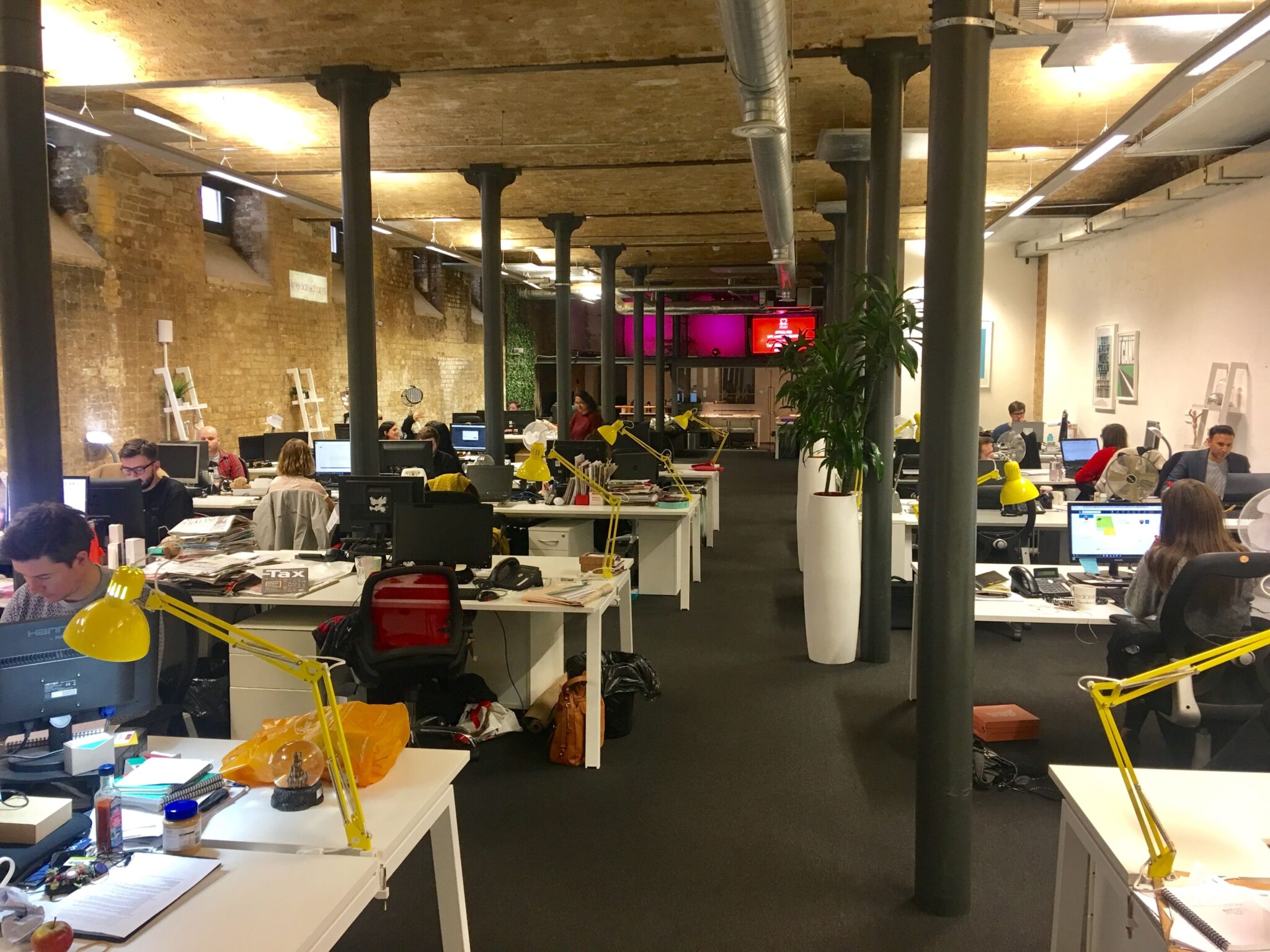 A busy open-plan 4media group office with brick ceilings, black columns and yellow desk lamps