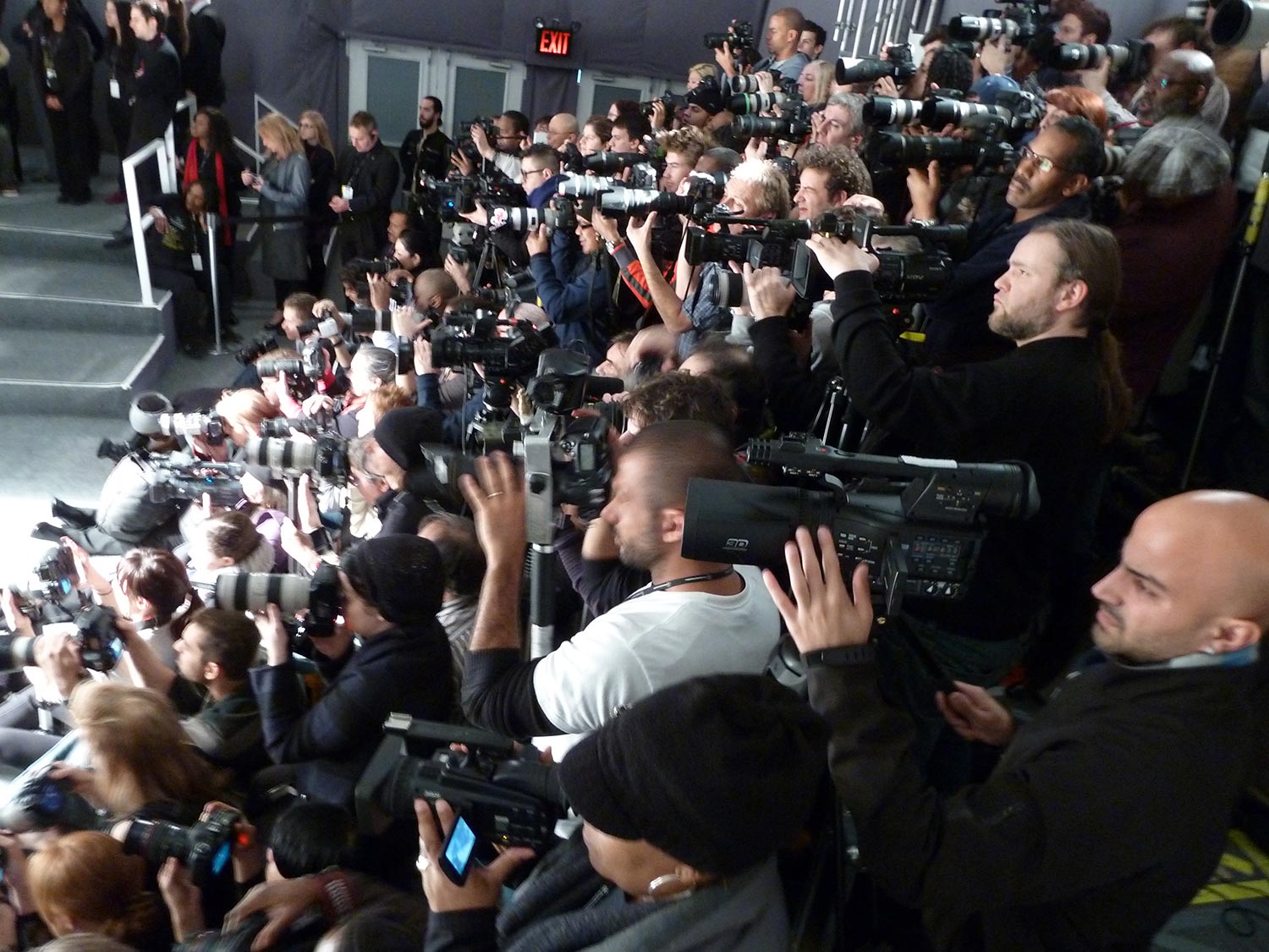 A dense group of press photographers and camera operators aiming toward an event stage
