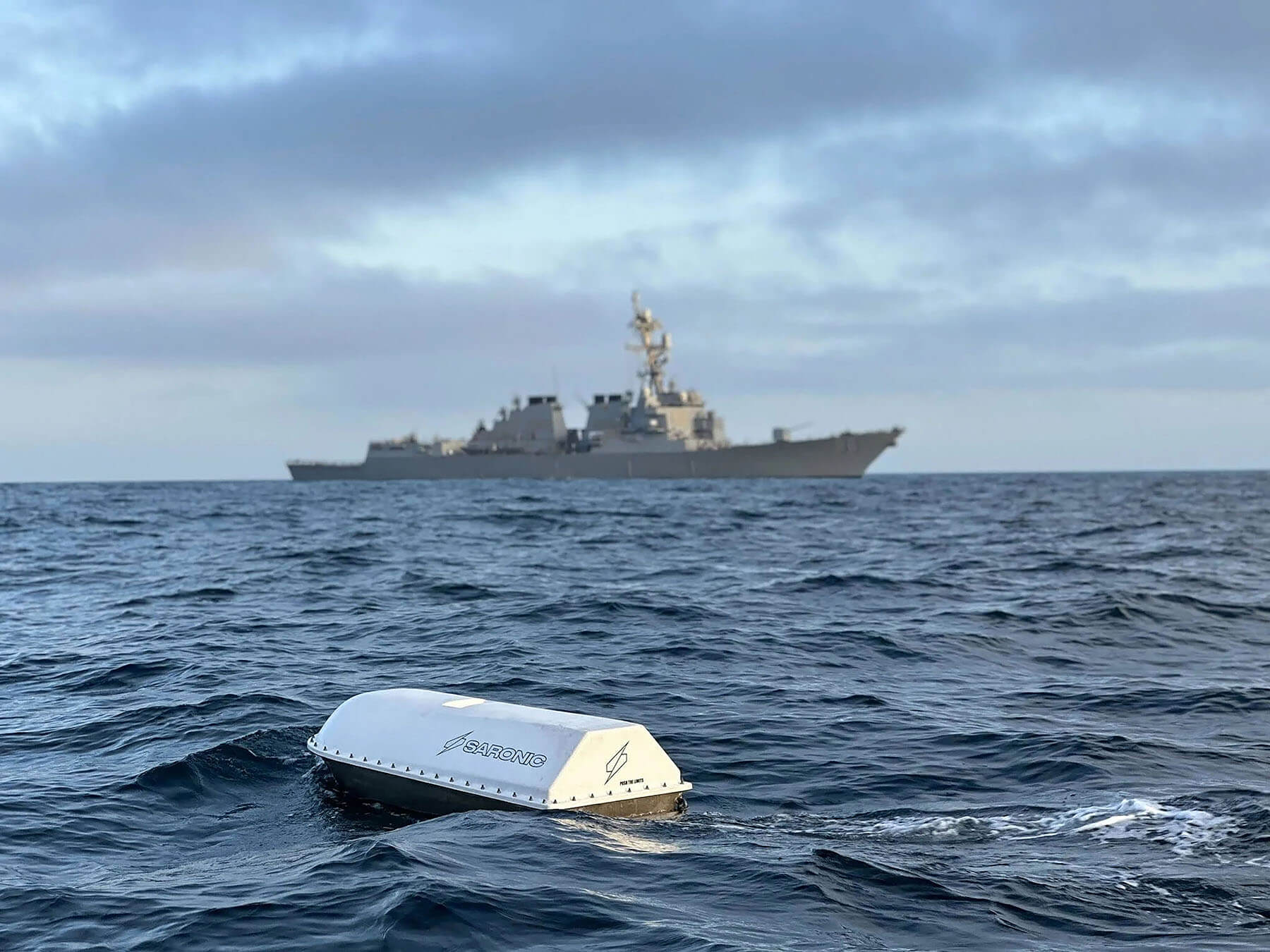 A small white Saronic autonomous vessel in the foreground with a naval ship behind it