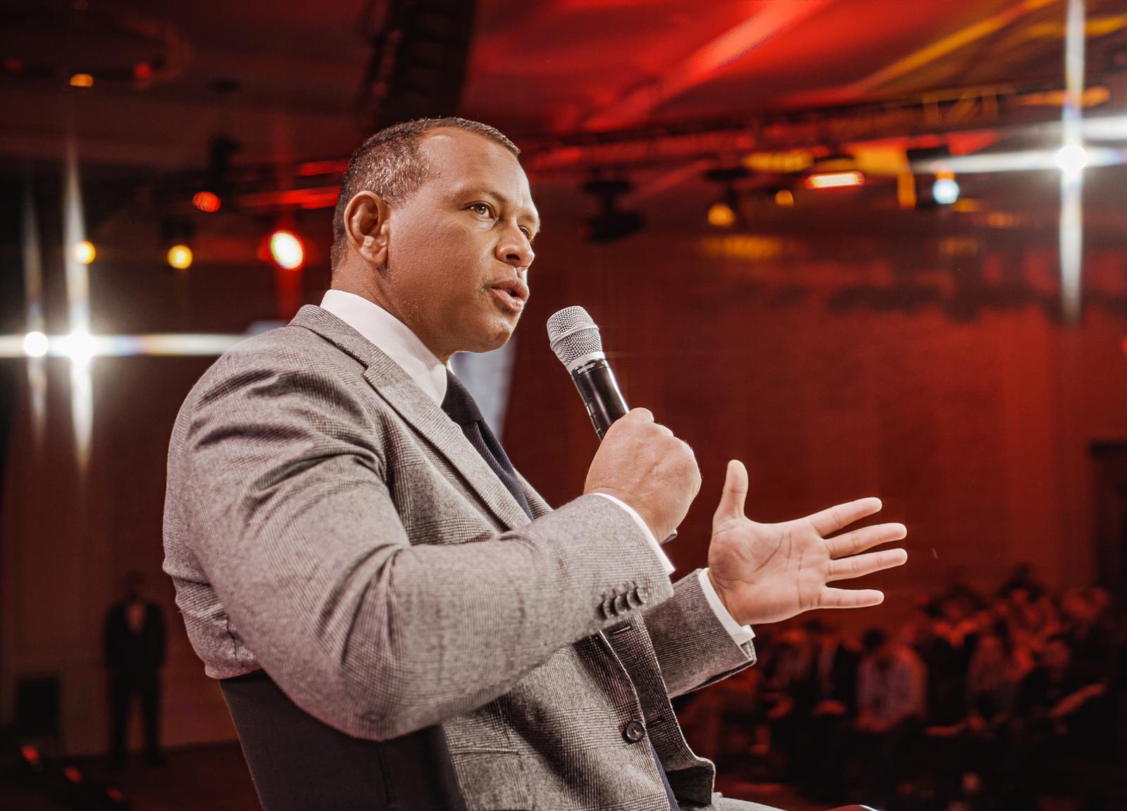 Alex Rodriguez in a gray suit, speaking into a handheld microphone
