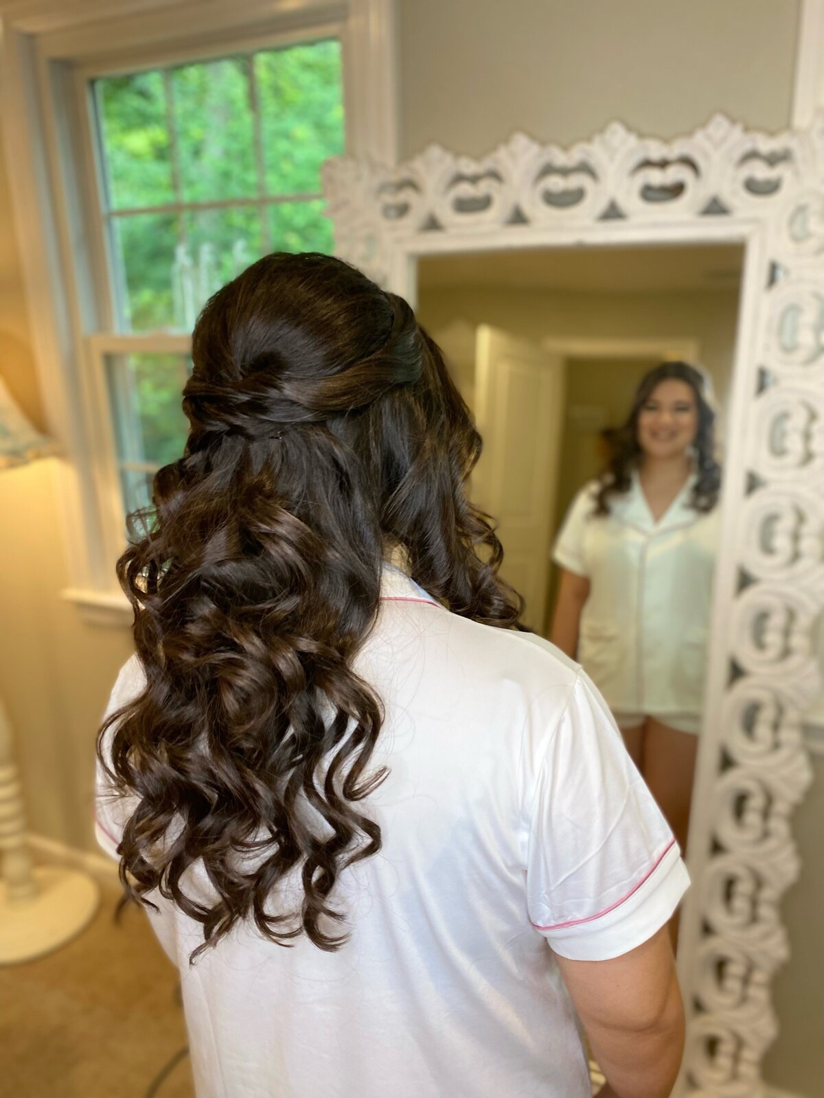 A bride views her curled half-up hairstyle in an ornate mirror