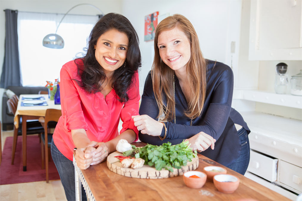 Aashi Vel, left, and Steph Lawrence leaning over a kitchen worktop with herbs and garlic