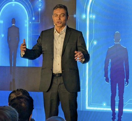 Aatish Dedhia speaking on a stage with a blue digital backdrop