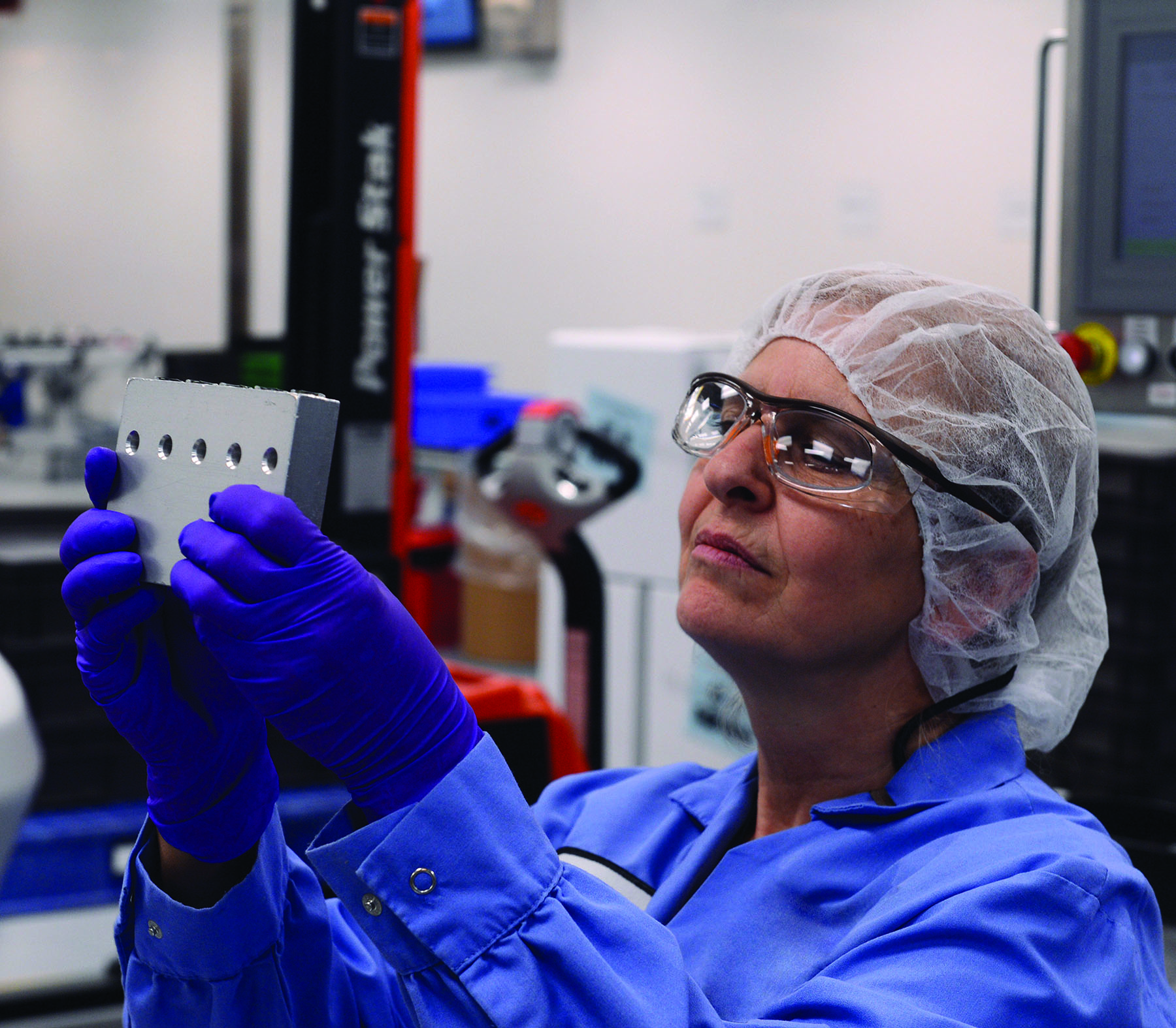 An AbbVie worker wearing protective glasses and blue gloves inspects a metal component