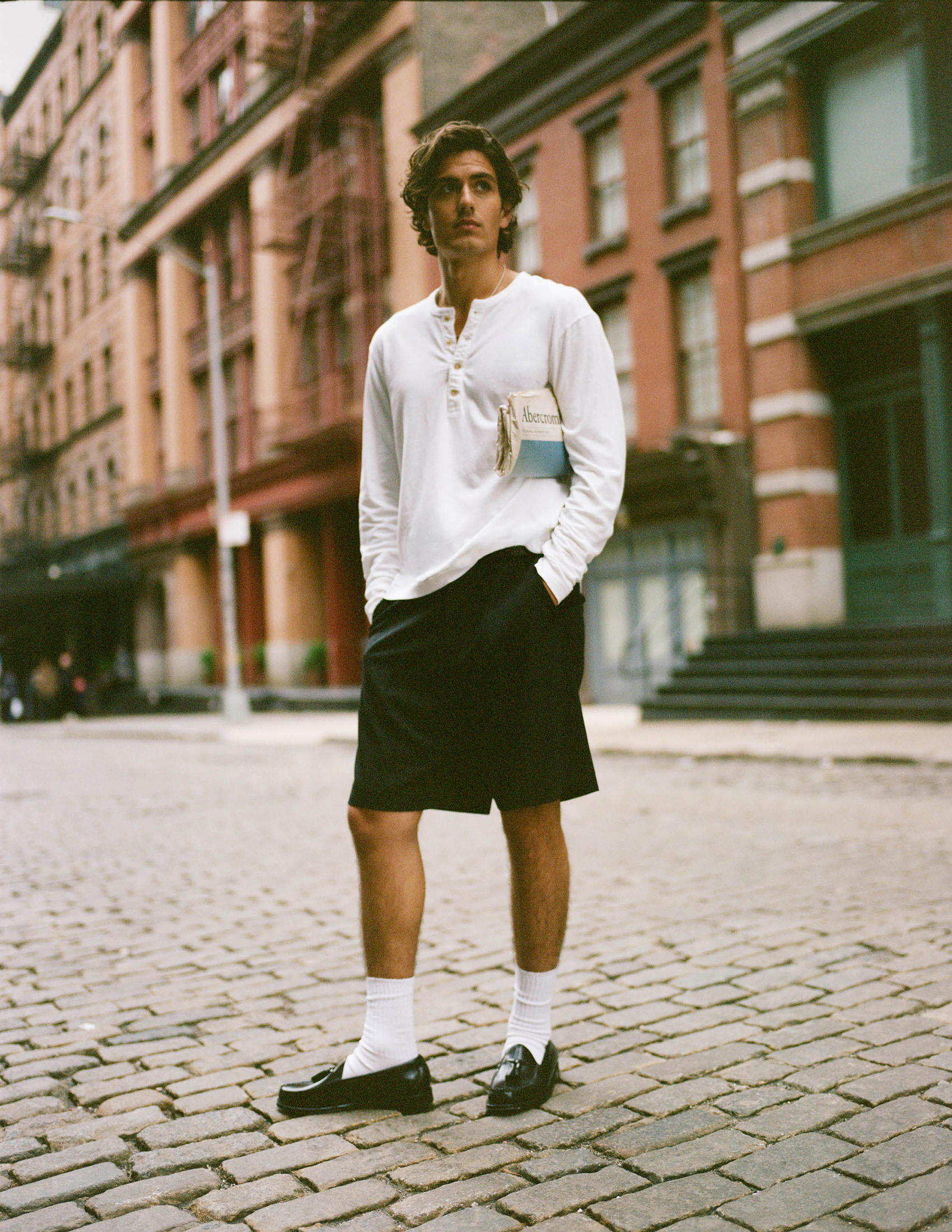 Abercrombie campaign model wearing a white Henley, black shorts and loafers on a cobbled city street