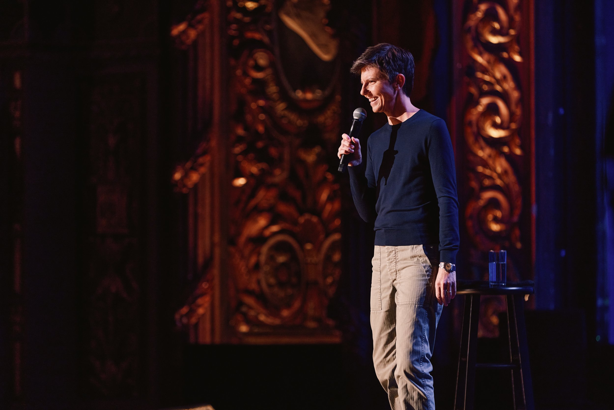Tig Notaro holding a microphone on an ornate theater stage