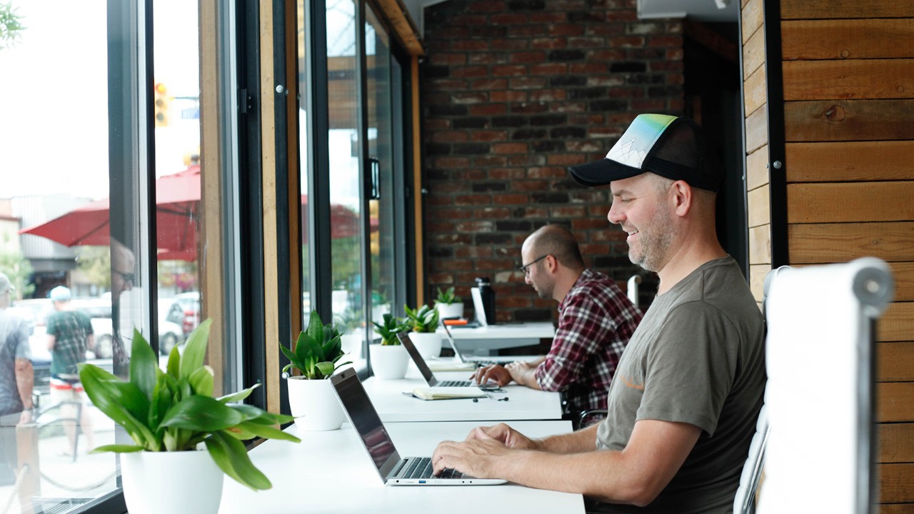 Two people working on laptops beside large windows in a collaborative workspace