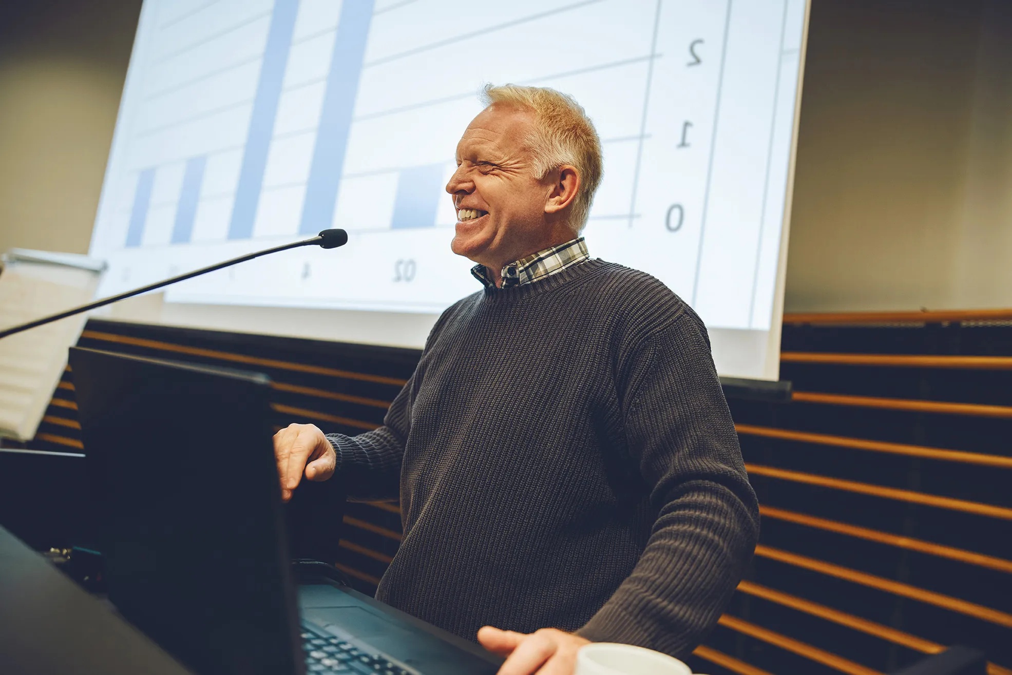 A presenter smiling at a microphone in front of financial charts during a seminar