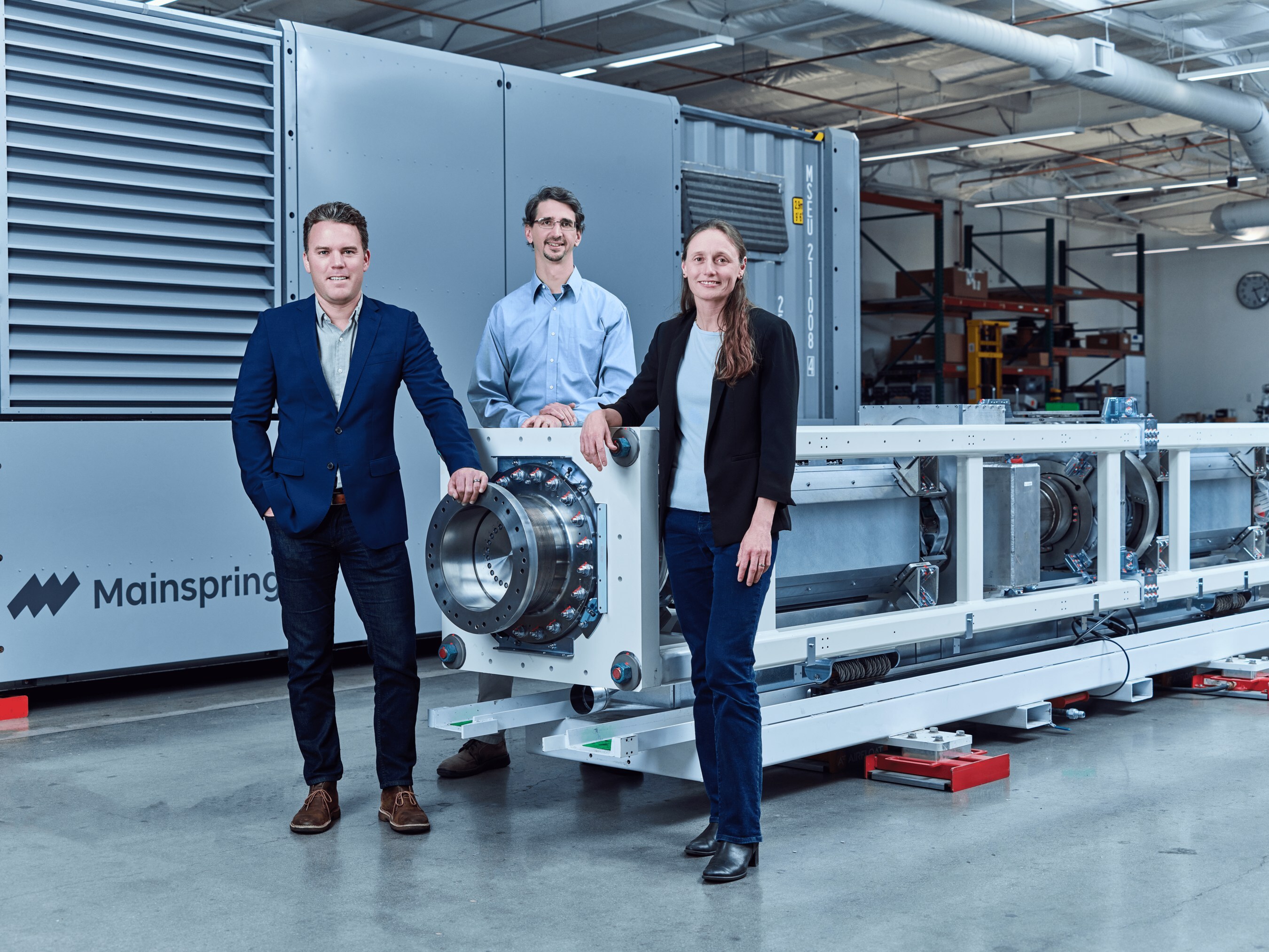 Adam Simpson, Matt Svrcek and Shannon Miller standing beside an exposed Mainspring linear generator in a manufacturing facility
