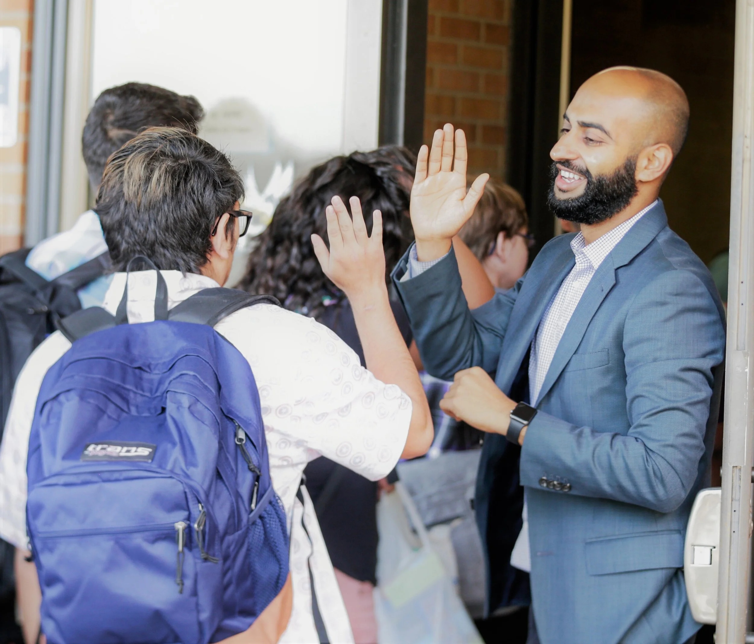 Adeel Khan smiles and raises his hand to high-five a student at a school entrance