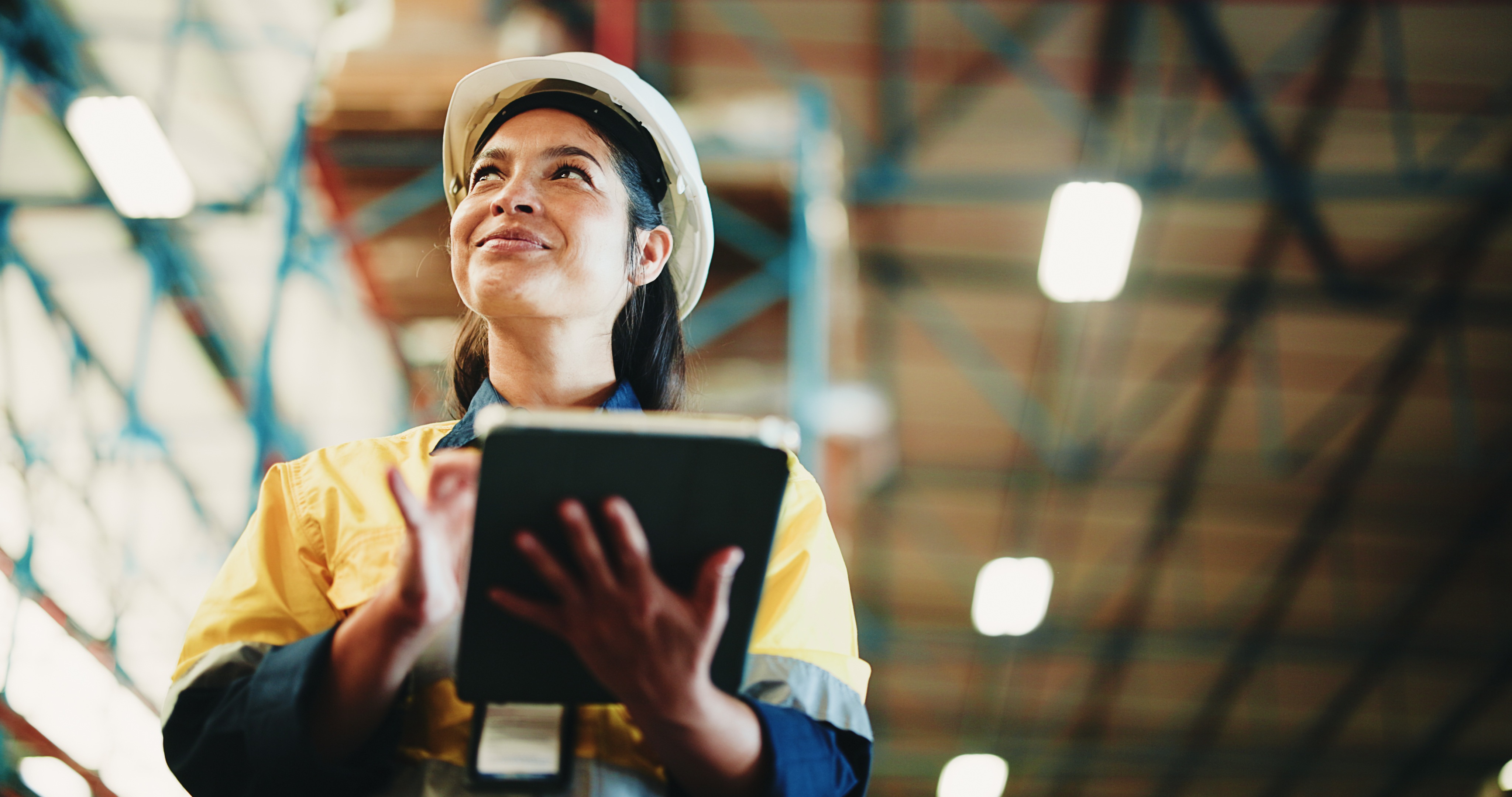 A warehouse worker in safety equipment holding a tablet among storage racks