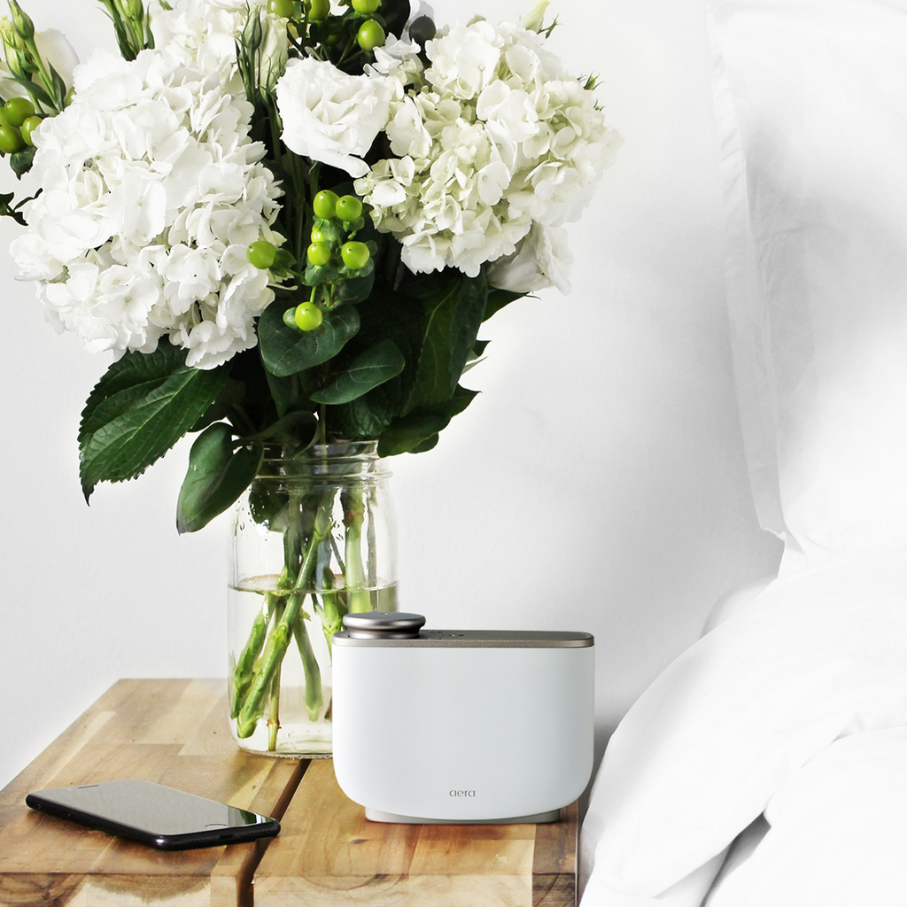 Aera diffuser beside a phone and white flowers on a bedside table