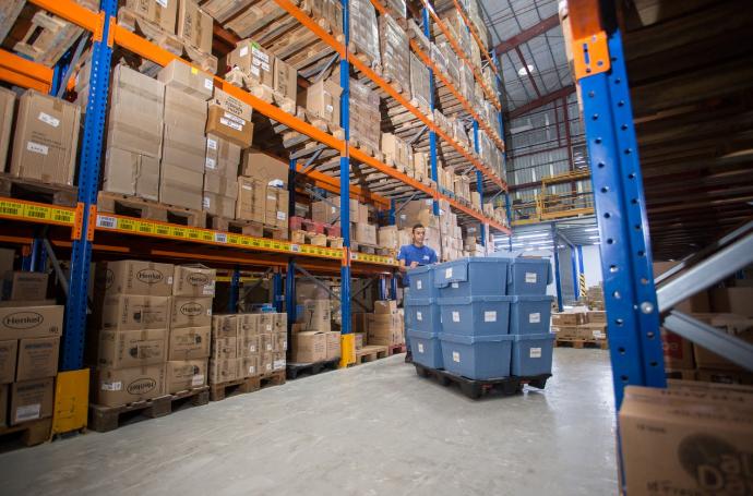 An employee moves blue bins between tall orange and blue warehouse racks filled with cartons.