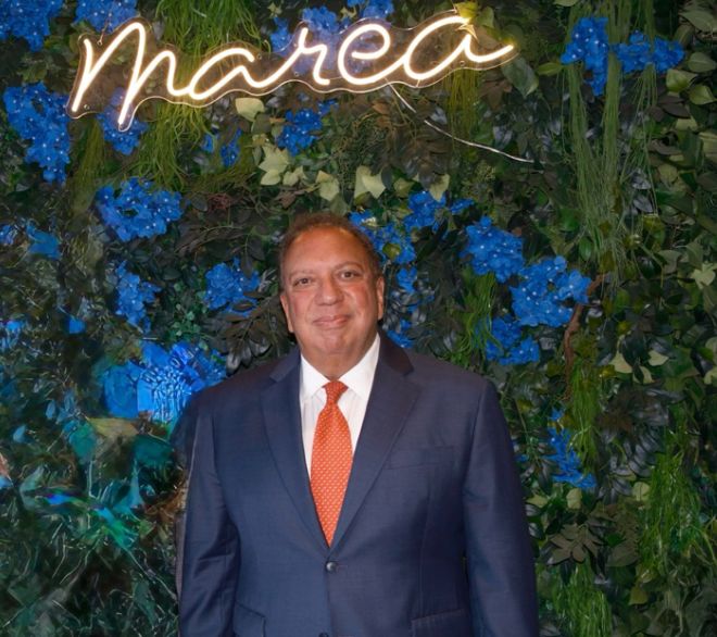 Ahmass Fakahany in a dark suit and orange tie in front of a floral wall and an illuminated Marea sign.