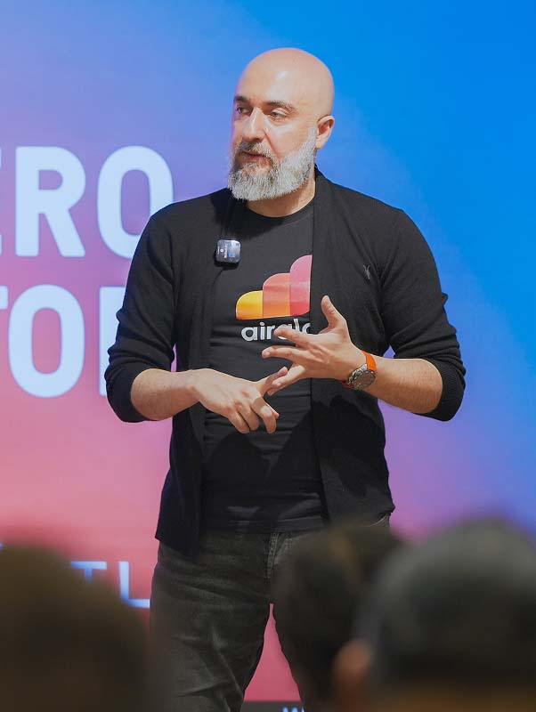 Özdemir speaking and gesturing on stage at an Antler event, wearing an Airalo shirt