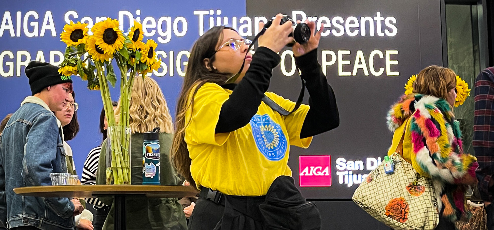 Attendees at an AIGA San Diego Tijuana gathering, with sunflowers and Designers for Peace signage