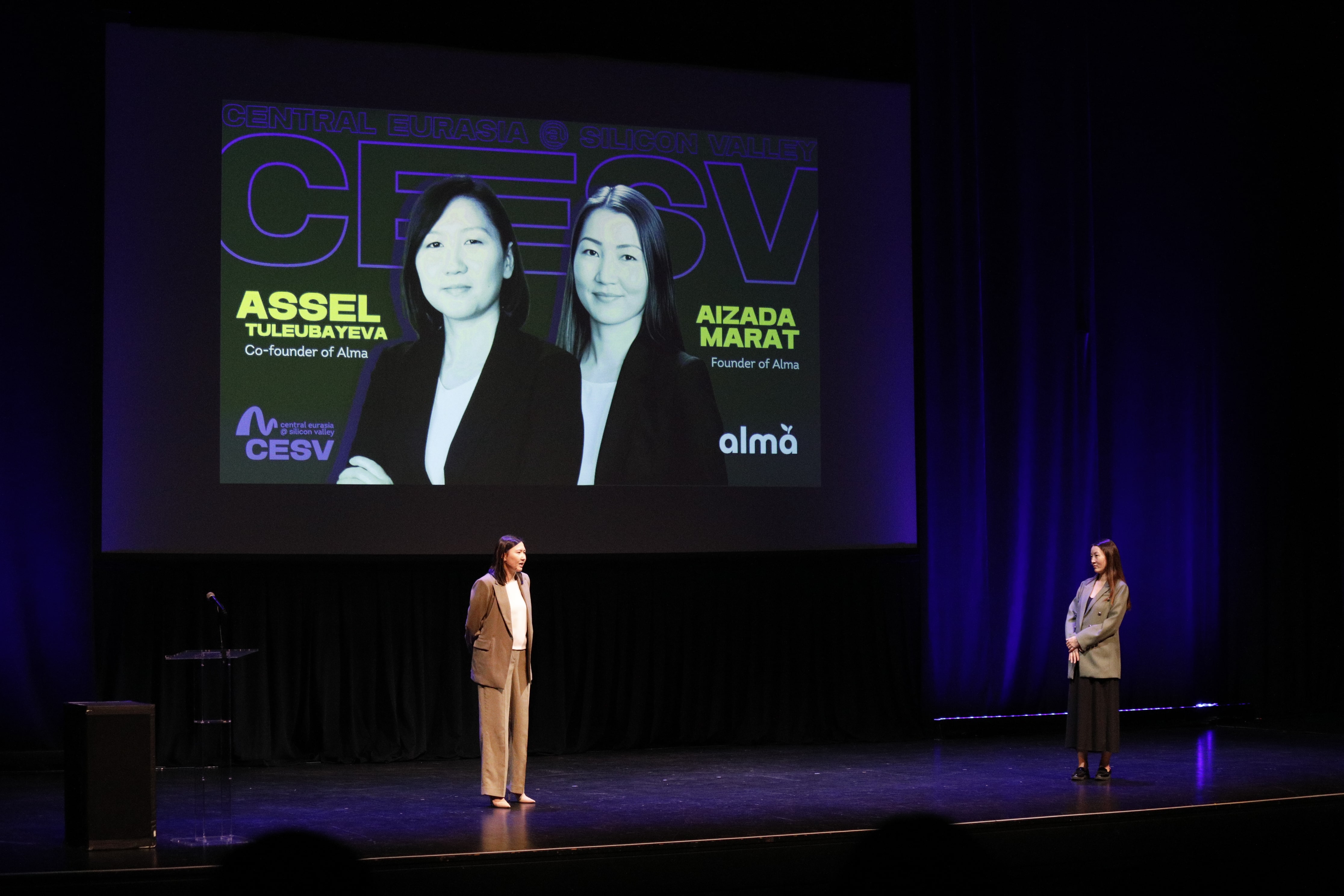 Assel Tuleubayeva and Aizada Marat standing onstage beneath a screen introducing them as Alma founders