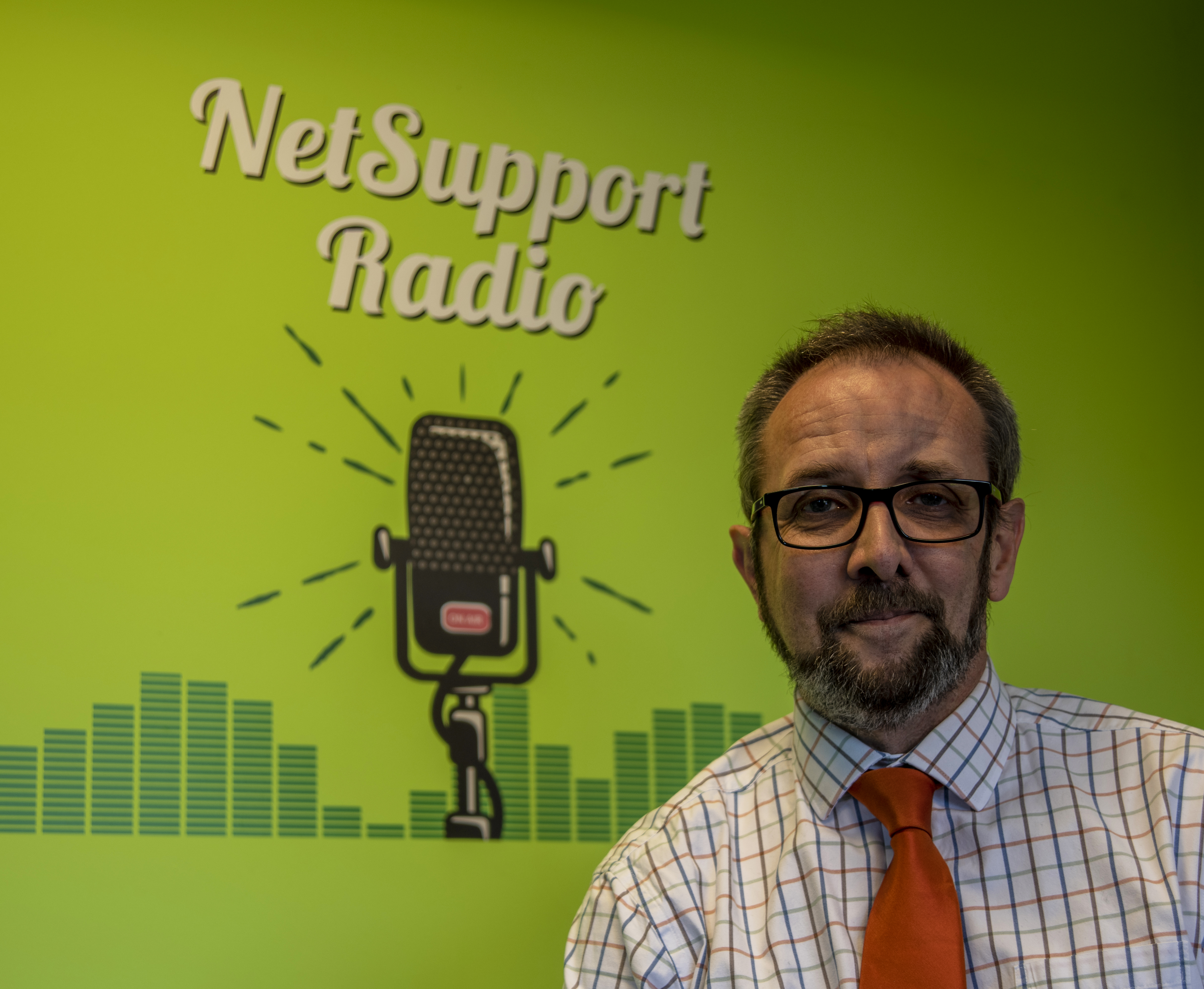 Al Kingsley standing in front of a green NetSupport Radio wall with a microphone illustration