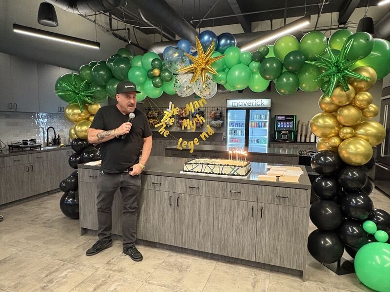 Alan Griefer holding a microphone beside a birthday cake beneath a balloon arch at Maverick’s office
