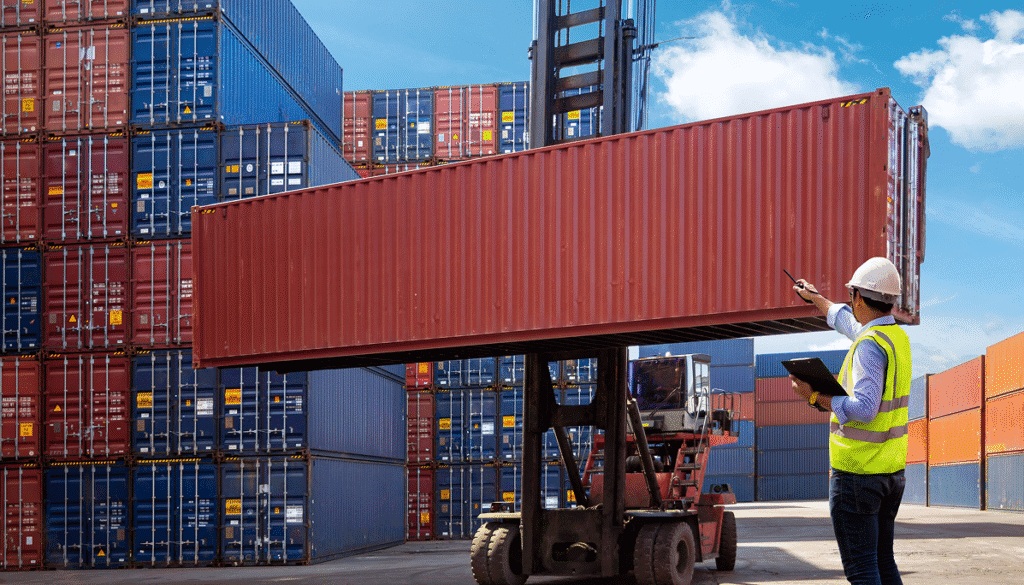 A container handler lifts a red shipping container while a worker directs the move