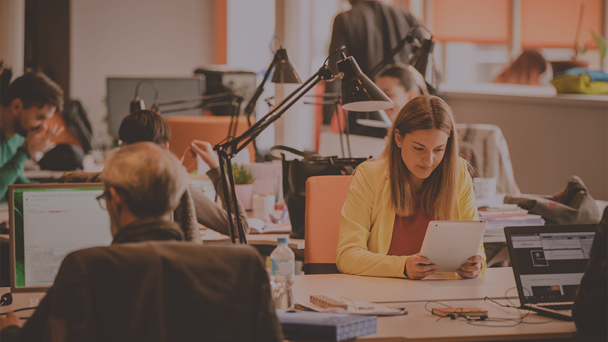 An office team works around desks while a woman reviews a tablet