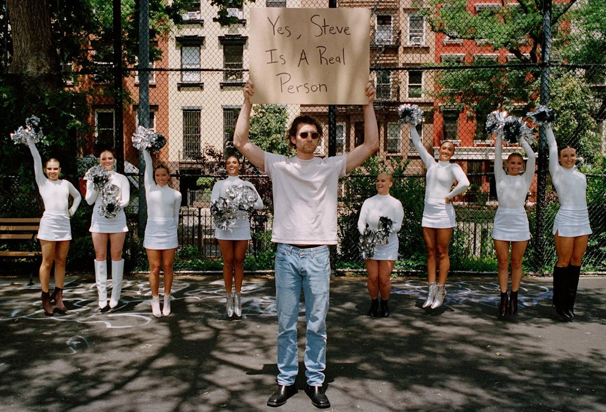 A man holding a handwritten sign reading Yes, Steve Is A Real Person on a New York basketball court, with cheerleaders behind him