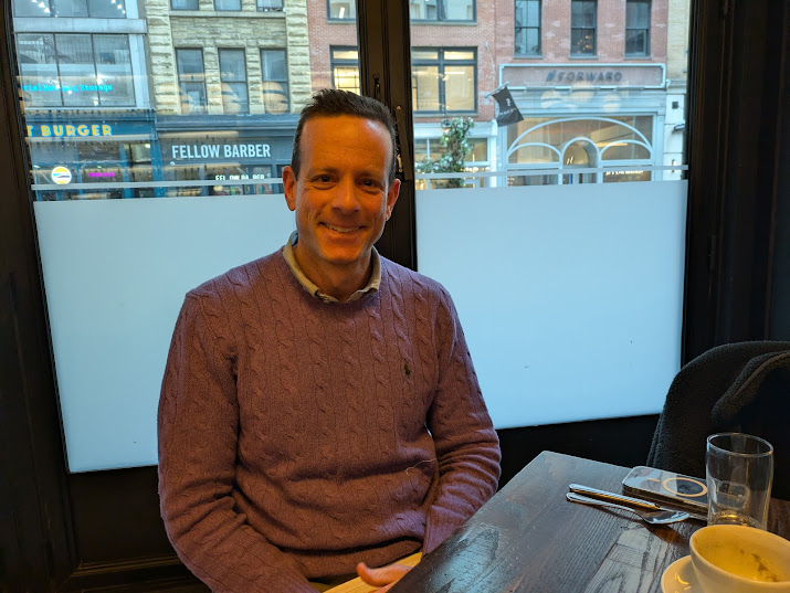 Alex Friedman seated beside a window at a restaurant table in Chelsea
