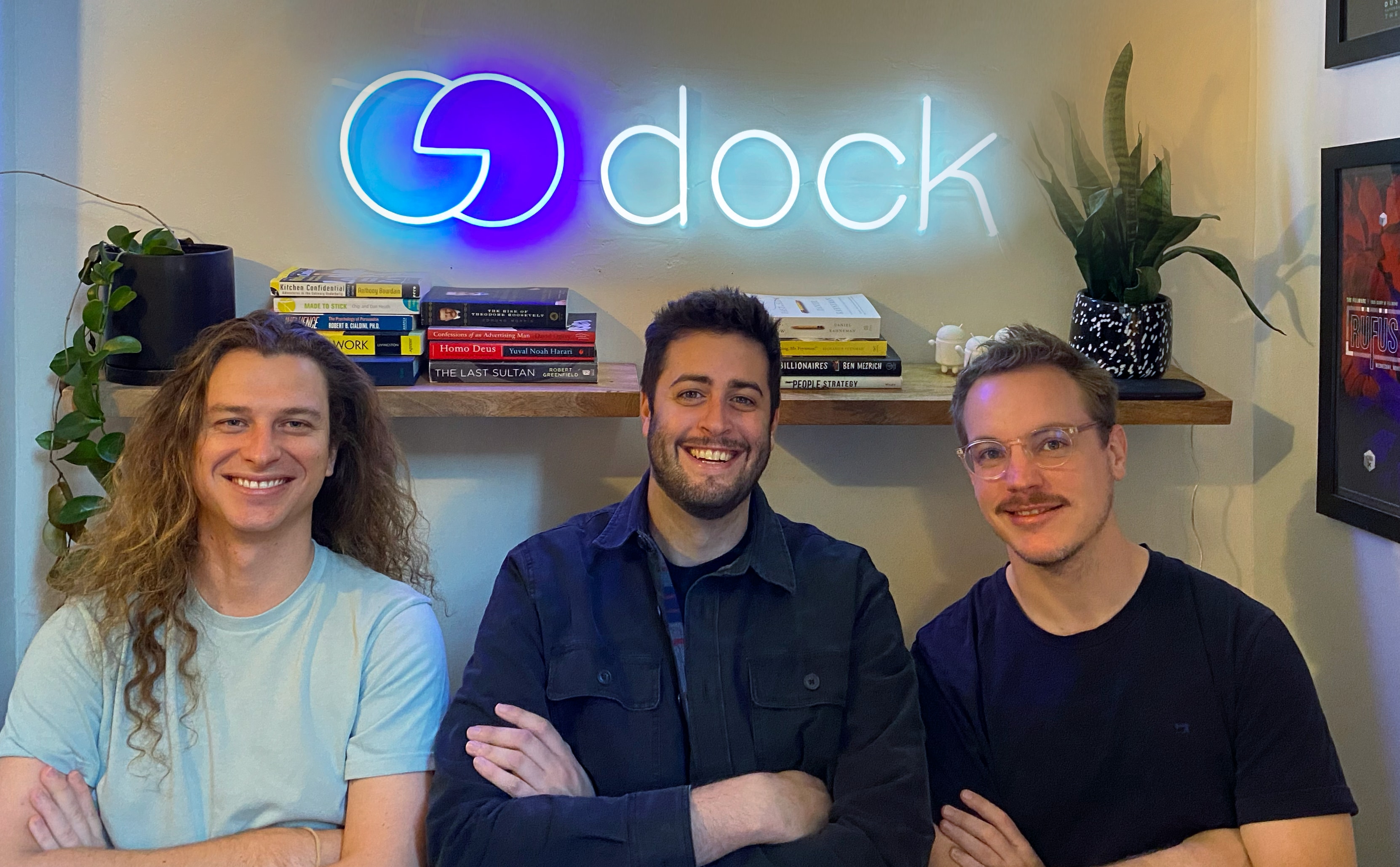 Dock’s three co-founders seated beneath a glowing Dock sign, with Alex Kracov in the center