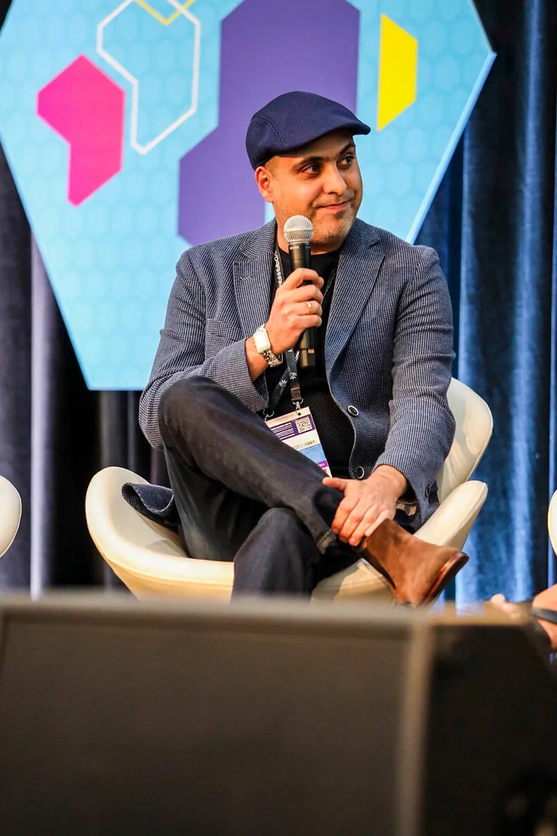 Alex Mojtahedi seated on stage, holding a microphone and wearing a dark cap and blazer