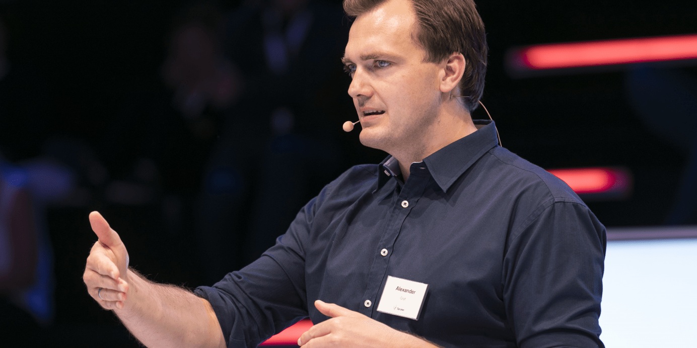 Alexander Graf speaking onstage with a headset microphone and gesturing with one hand