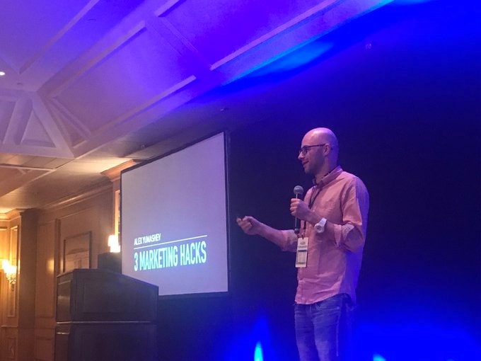 Alex Yumashev speaking onstage beside a slide titled 3 Marketing Hacks