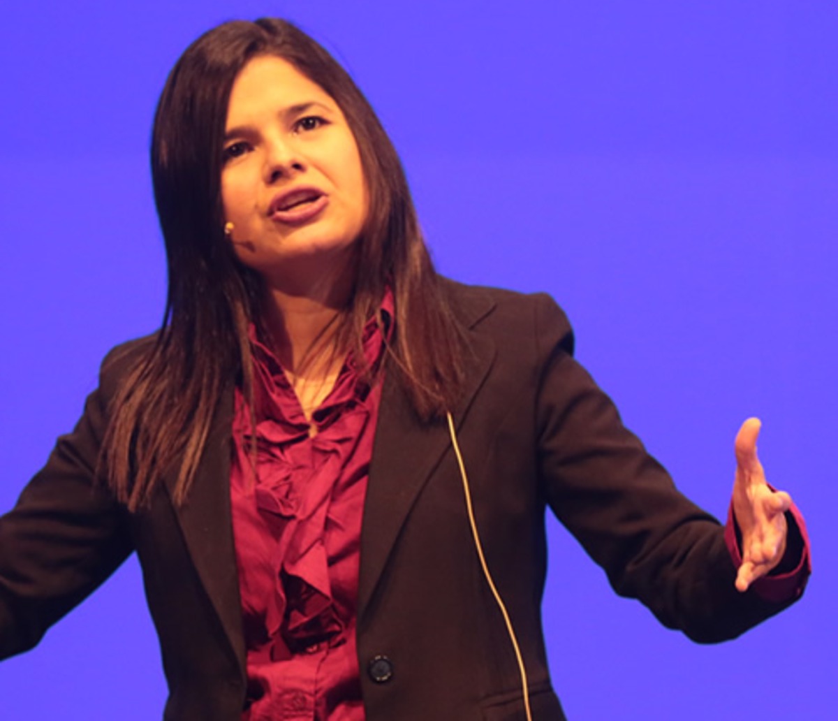 Aleyda Solís speaking on stage in a dark jacket and red blouse against a blue backdrop