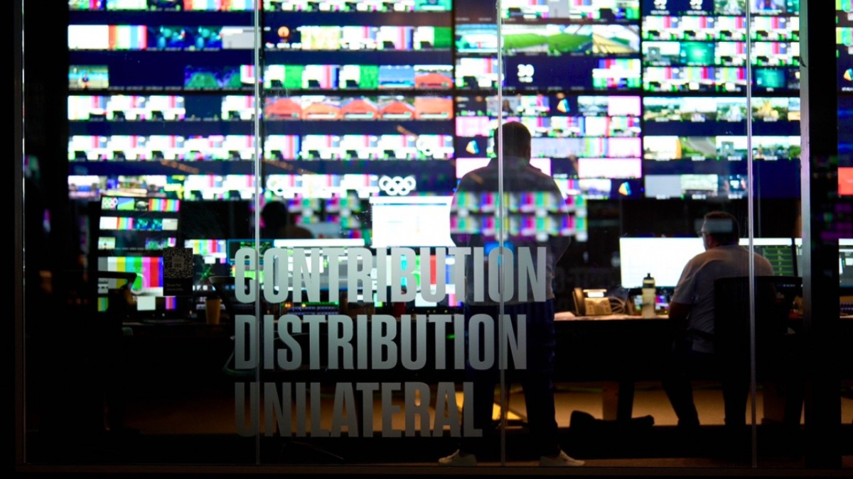 Operators at desks facing a wall of Olympic broadcast monitoring screens