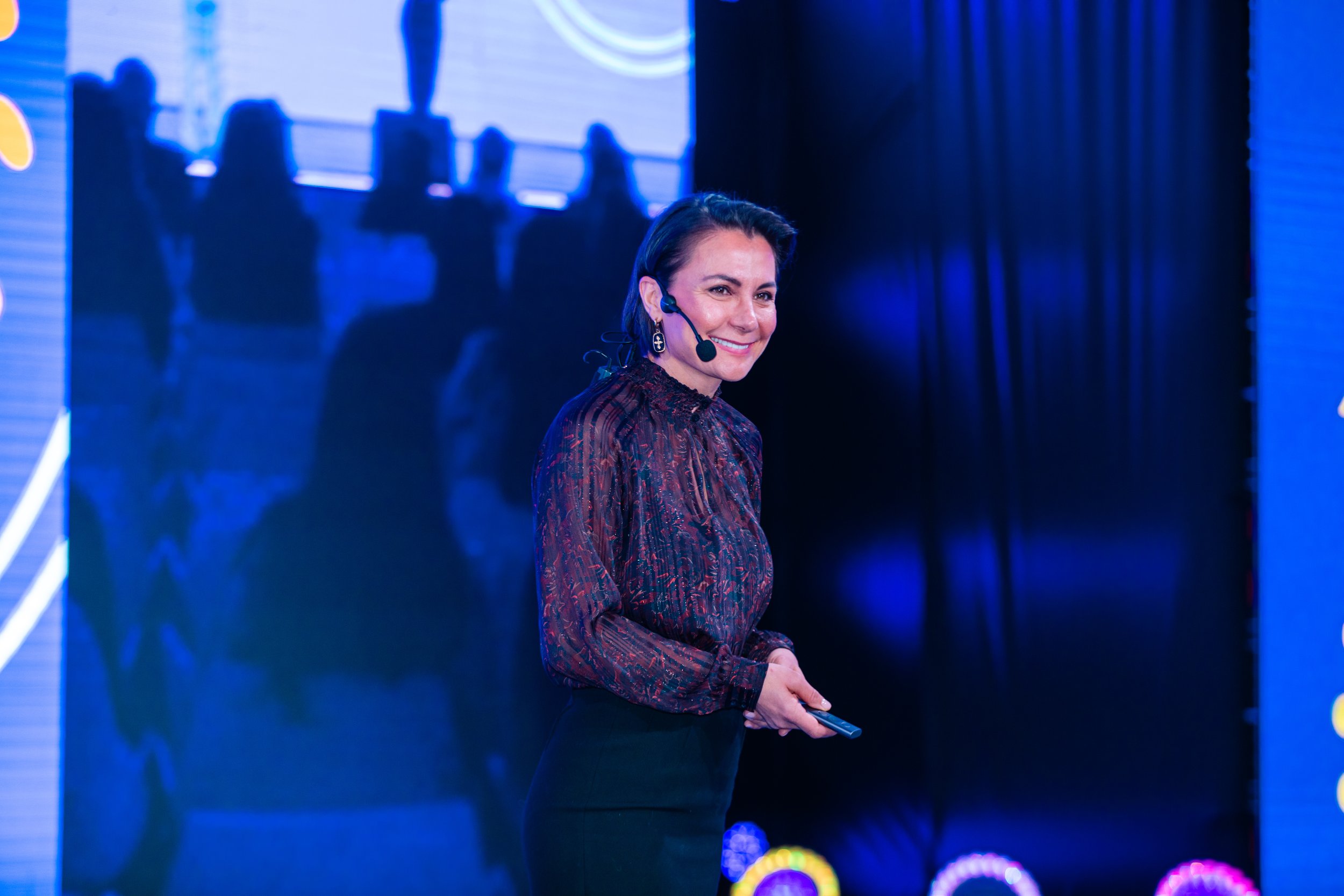 Alla Idrisova speaking on a blue-lit stage with a headset microphone and presentation remote