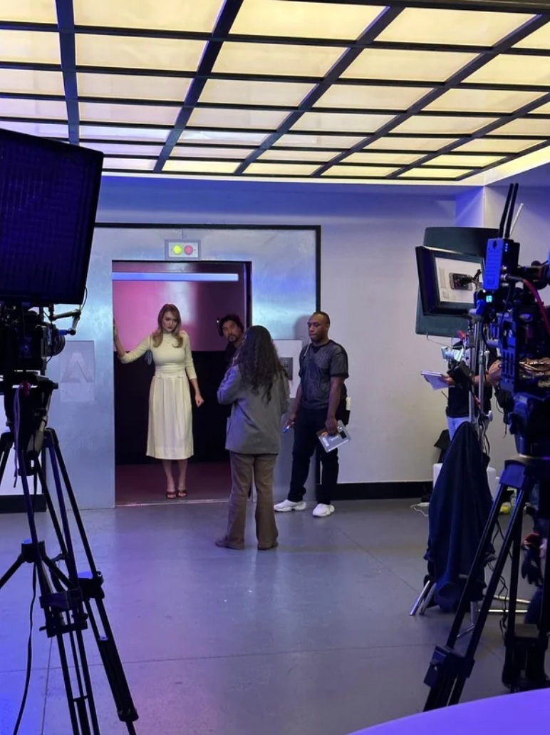 Behind-the-scenes production photograph with actors near an elevator, crew members, cameras and lighting equipment