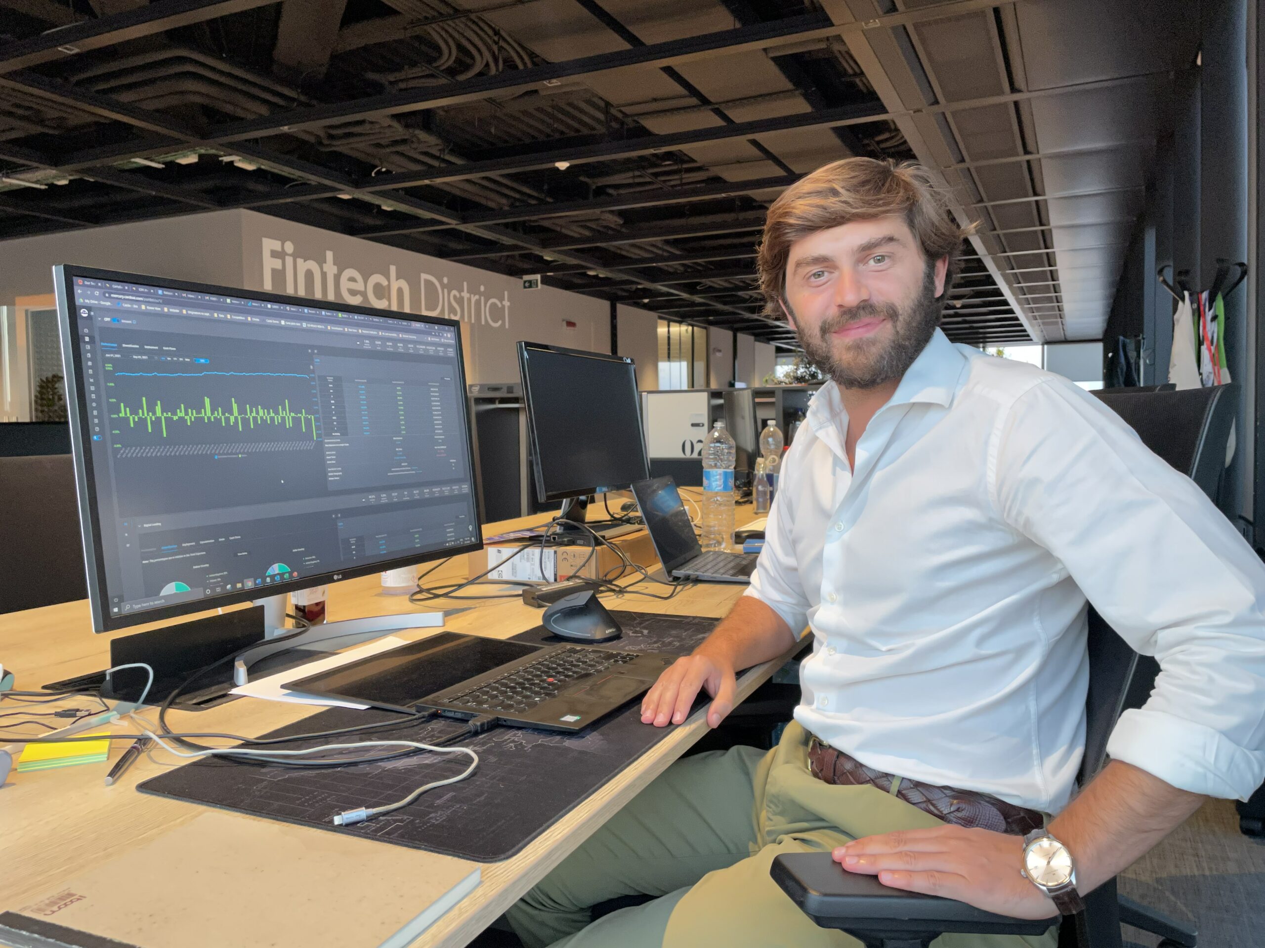 Altin Kadareja seated beside a large monitor displaying financial analytics at the Fintech District workspace