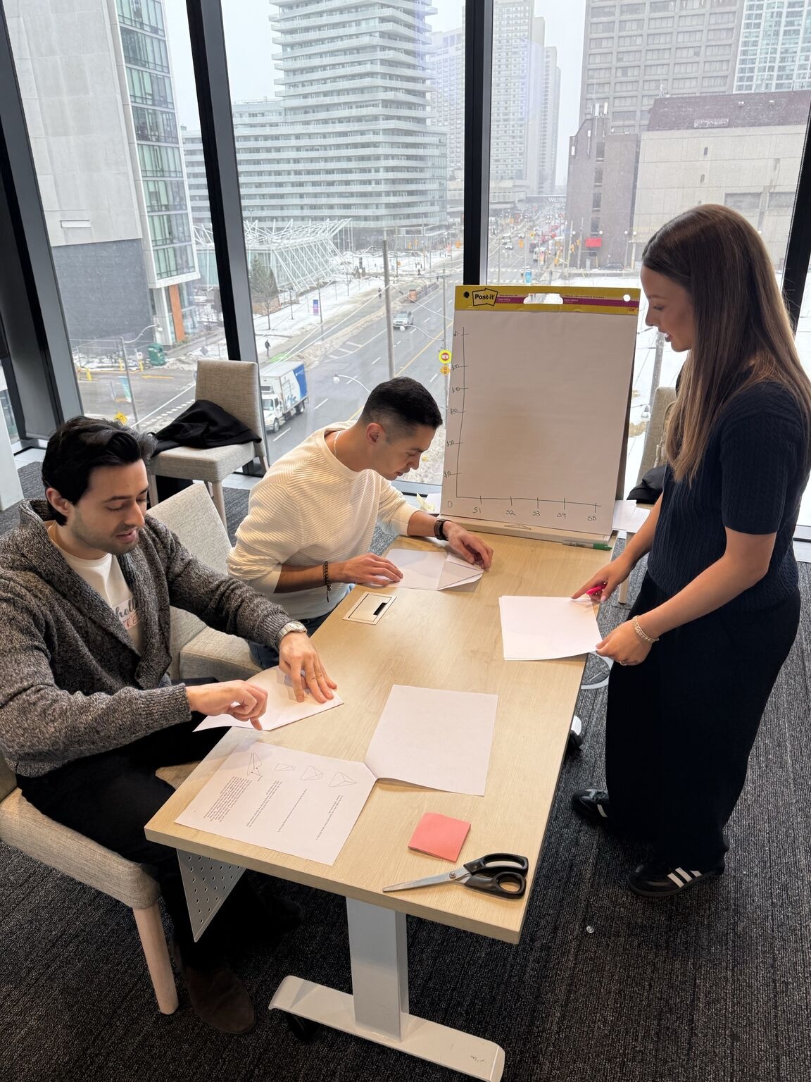Three Tru colleagues folding paper airplanes around a table beside a flip chart at a Canadian team offsite