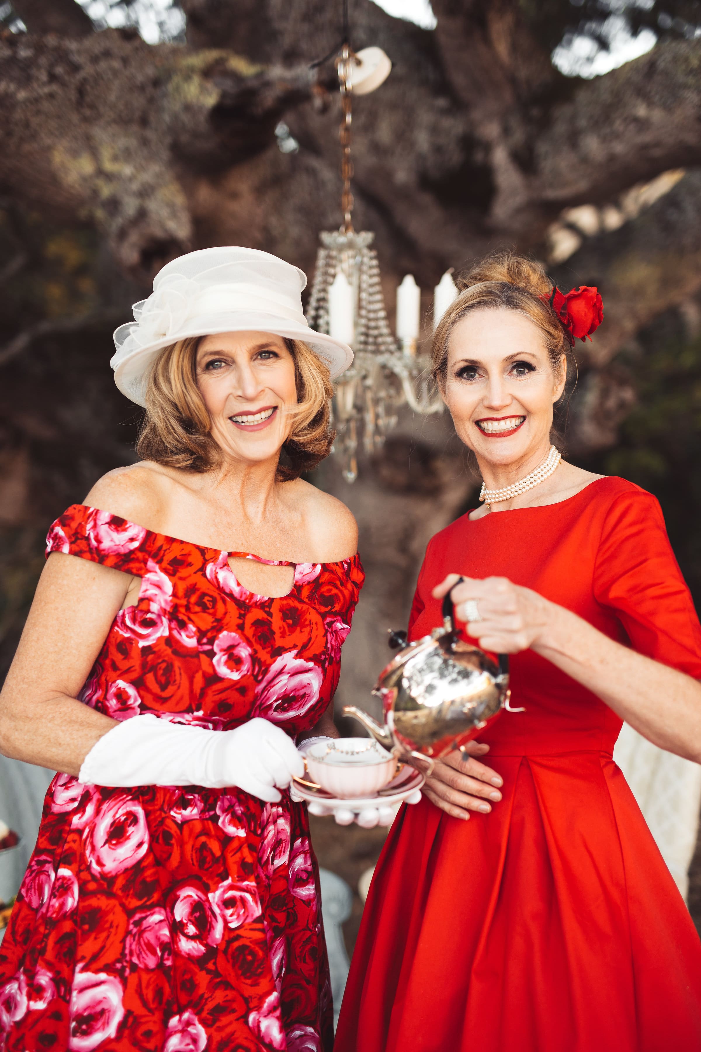 Jennifer Chapin in a floral dress and white hat beside Amanda Jones in red, holding a silver teapot.