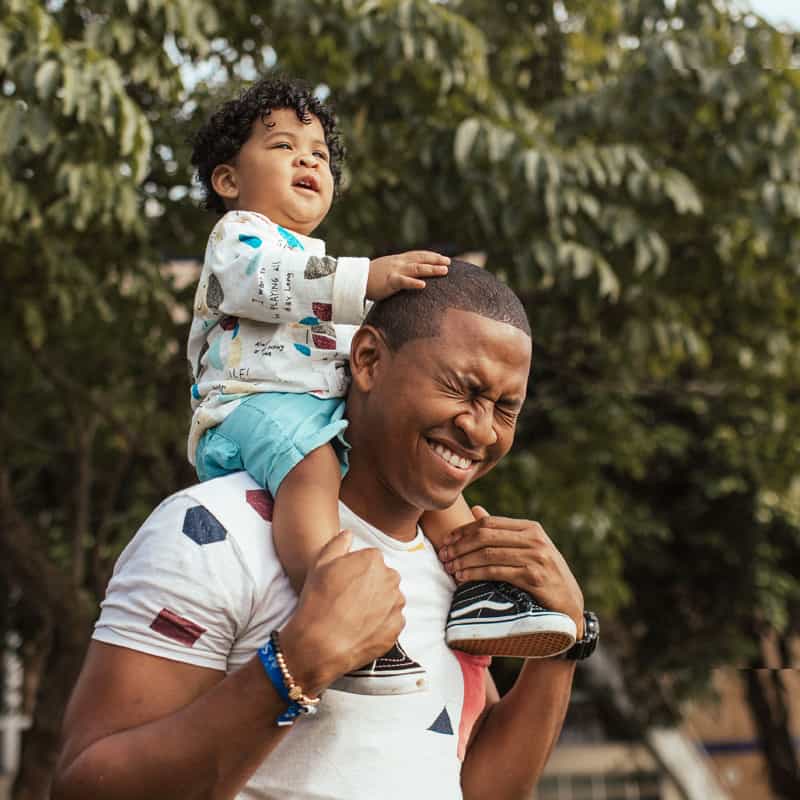 A smiling father carrying his young child on his shoulders in a Dear Dad Tour campaign image