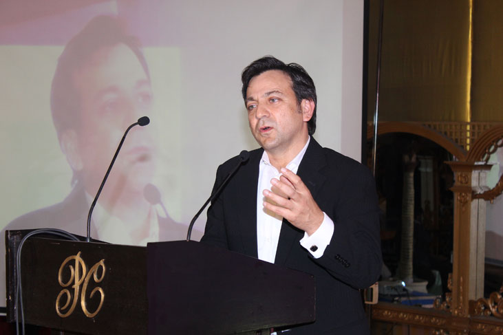 Amir Wain speaking at a lectern with a microphone, a projected portrait behind him
