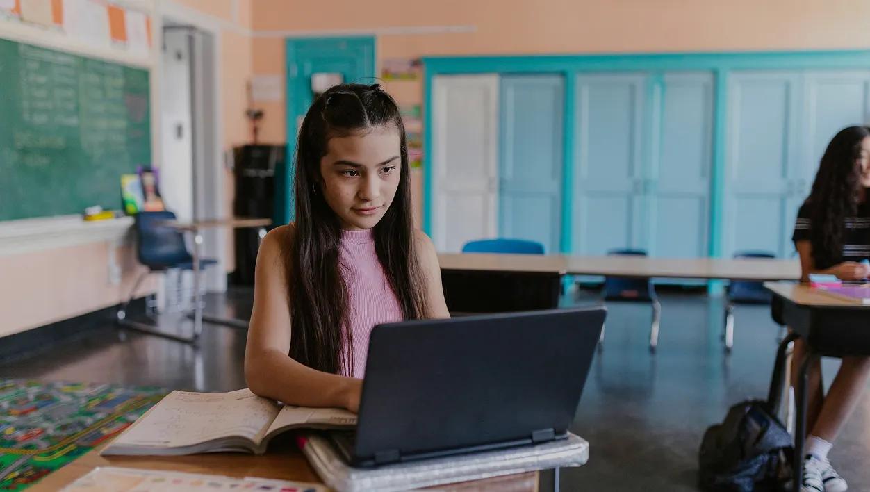A student sits at a classroom desk with a laptop and an open book.