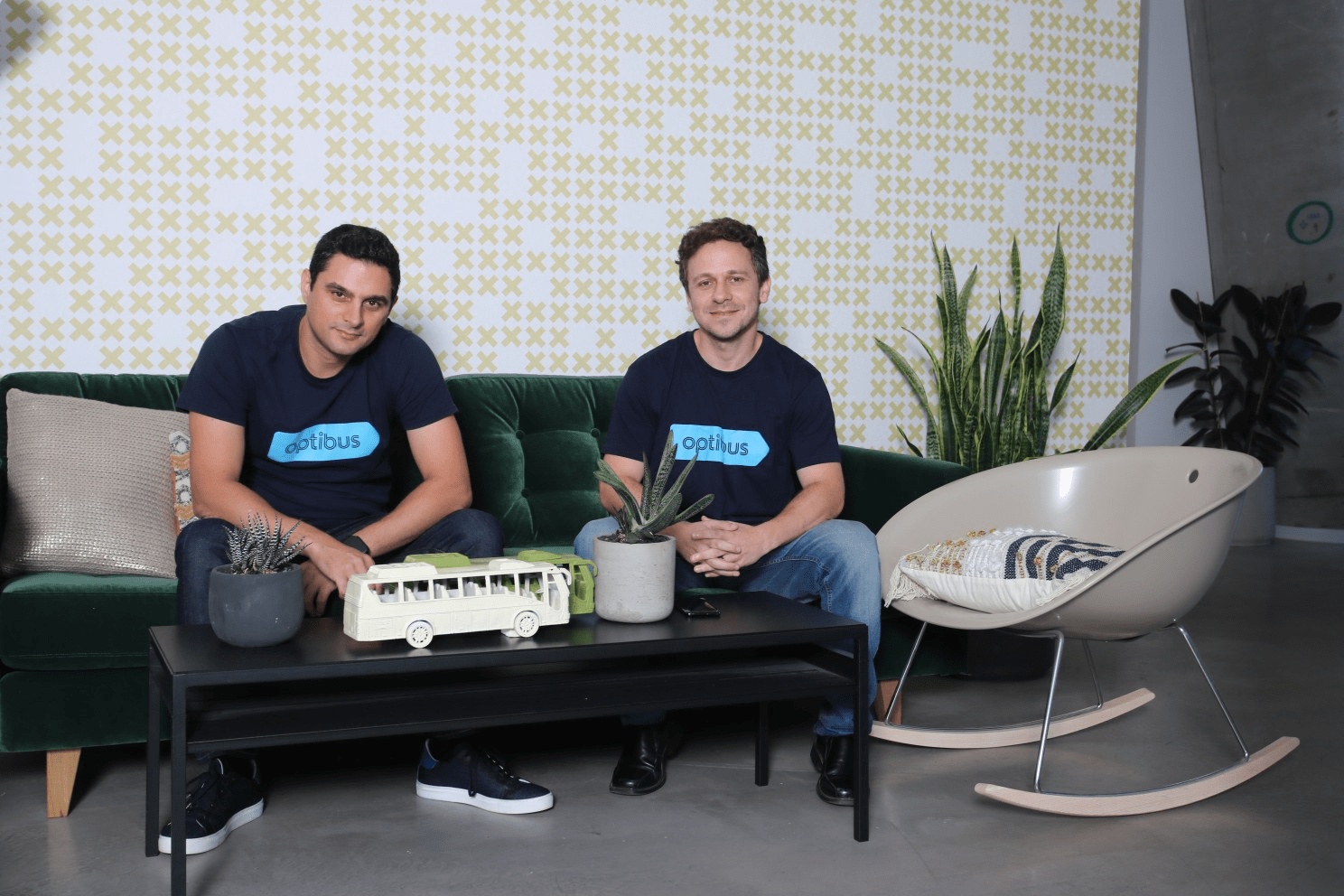 Amos Haggiag, left, and co-founder Eitan Yanovsky seated together in Optibus shirts, with a model bus on the table.