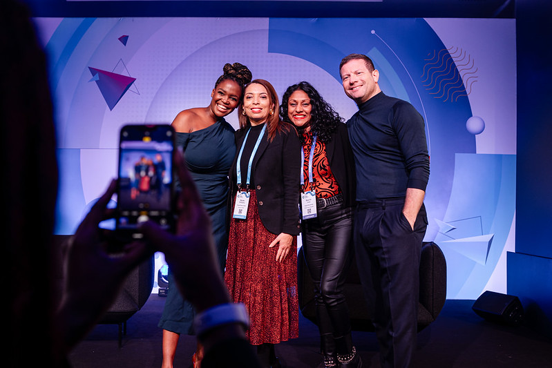 Four event speakers pose together on a lit stage while an attendee photographs them