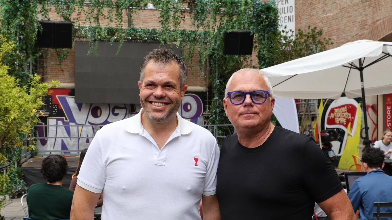 Andrea Magelli in a white polo shirt beside Gianluca Cristoni, with greenery and an outdoor stage behind them