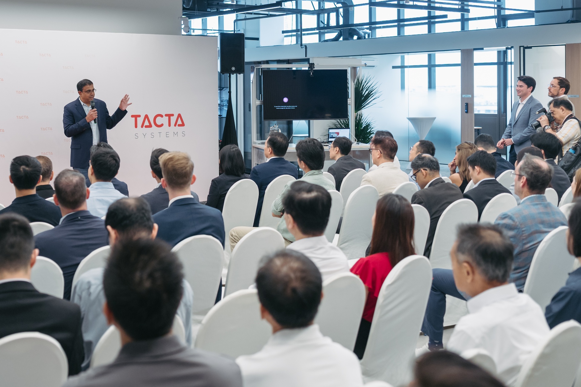 An audience attends an event with a speaker beside Tacta Systems branding