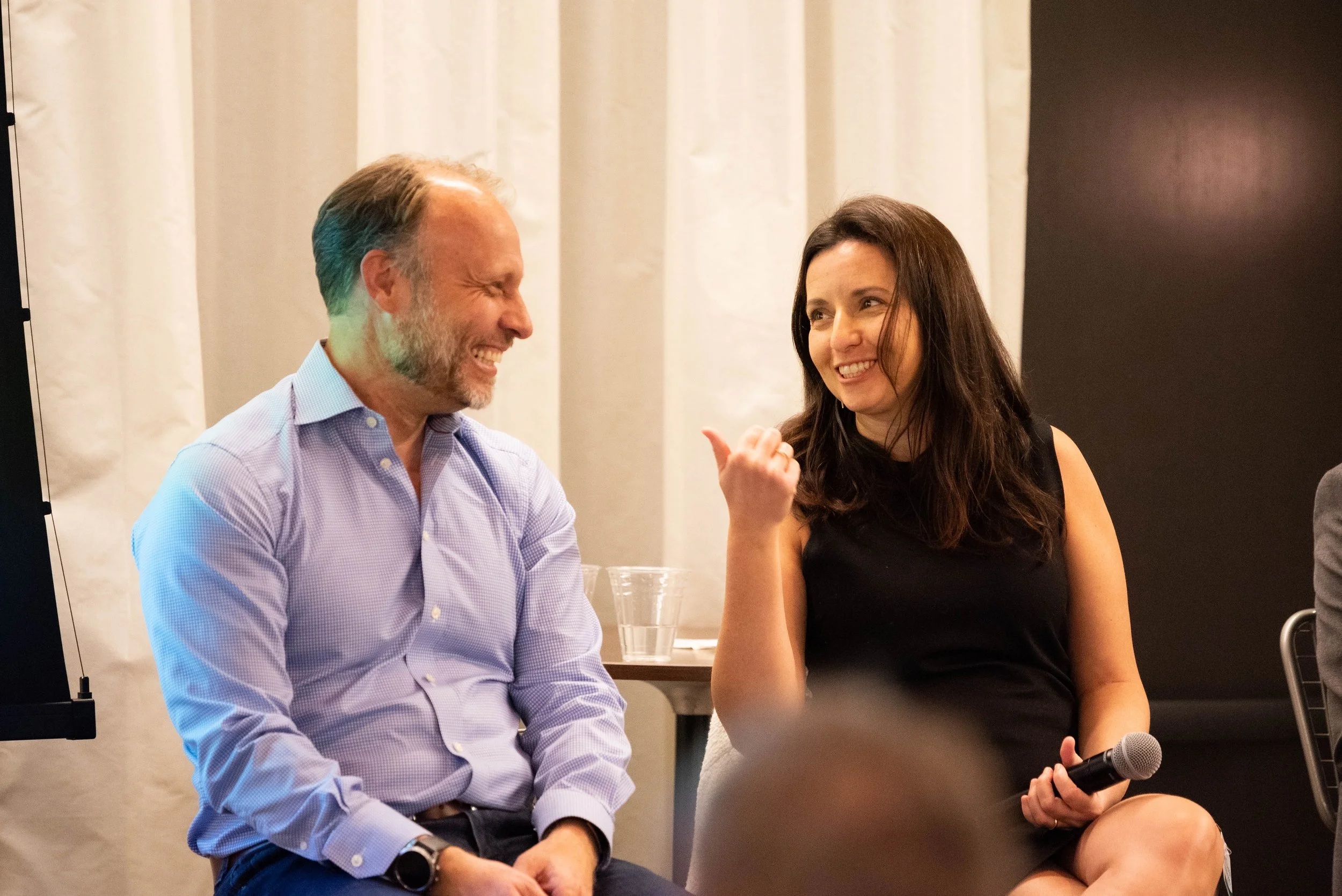 Andreas Roell, at left, laughing during an event conversation beside a woman holding a microphone.