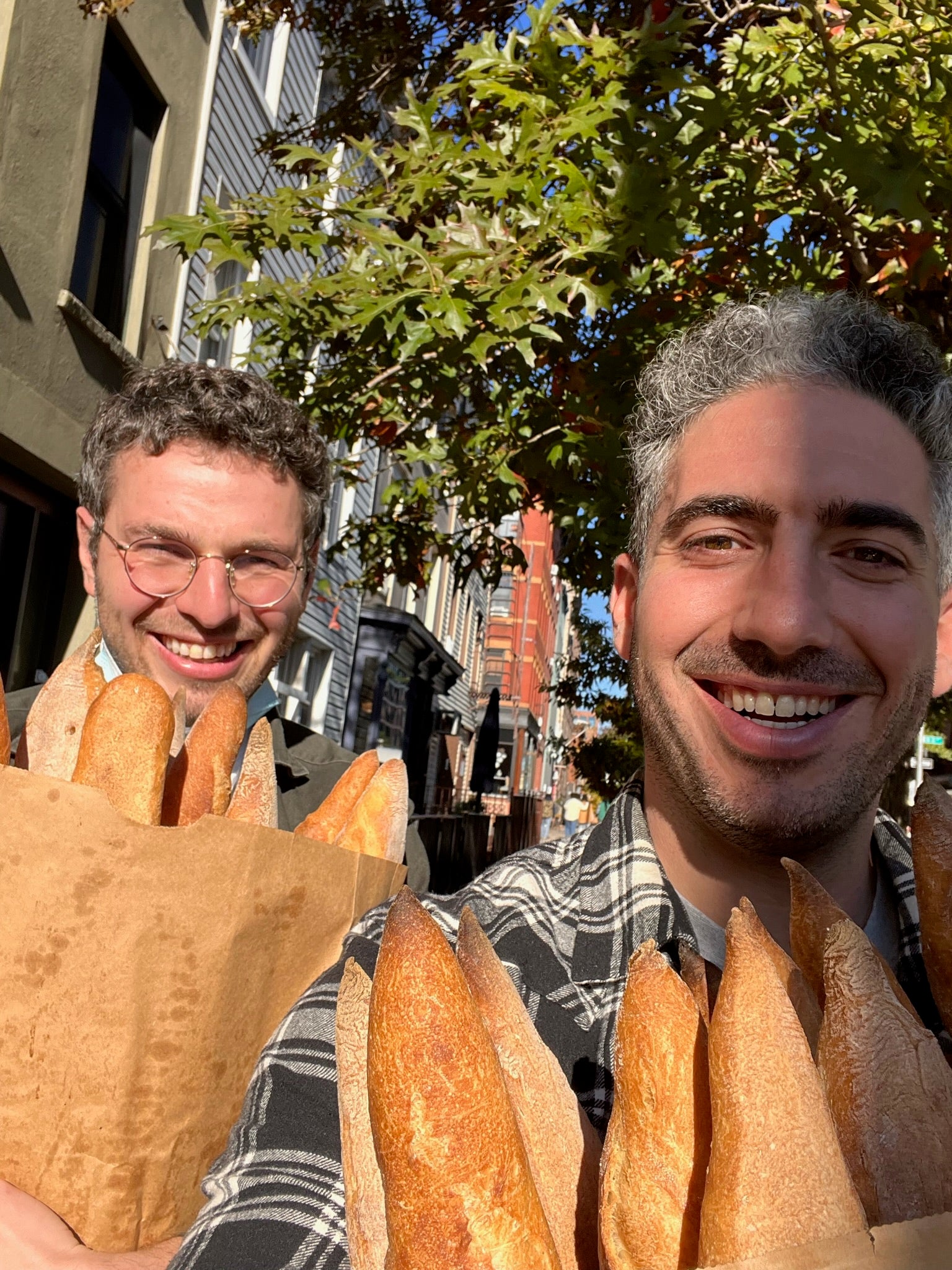 Andrew Benin, at left, and a companion smiling outdoors while carrying bags of baguettes